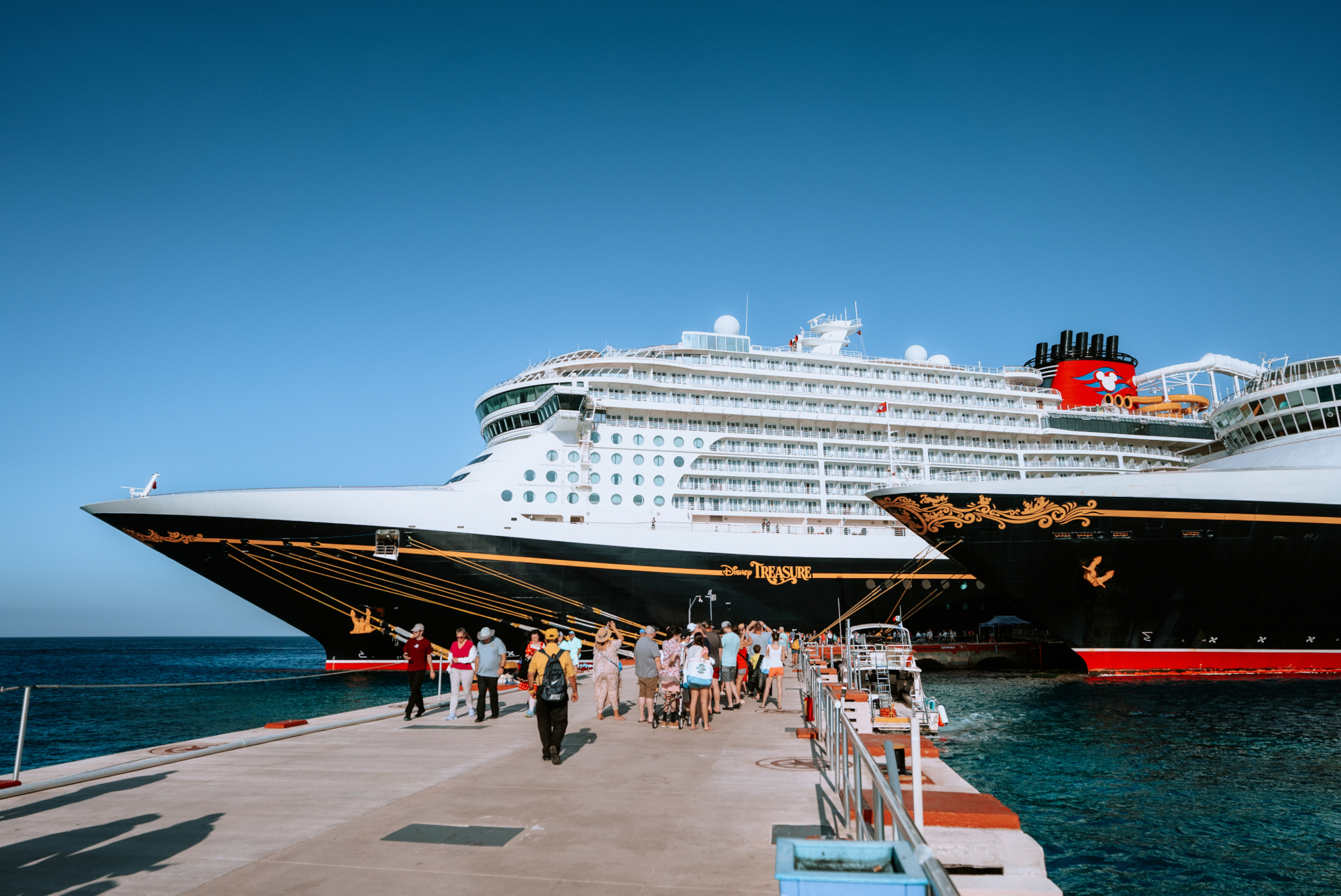 A Disney cruise ship is docked beside a pier with guests walking toward the ship