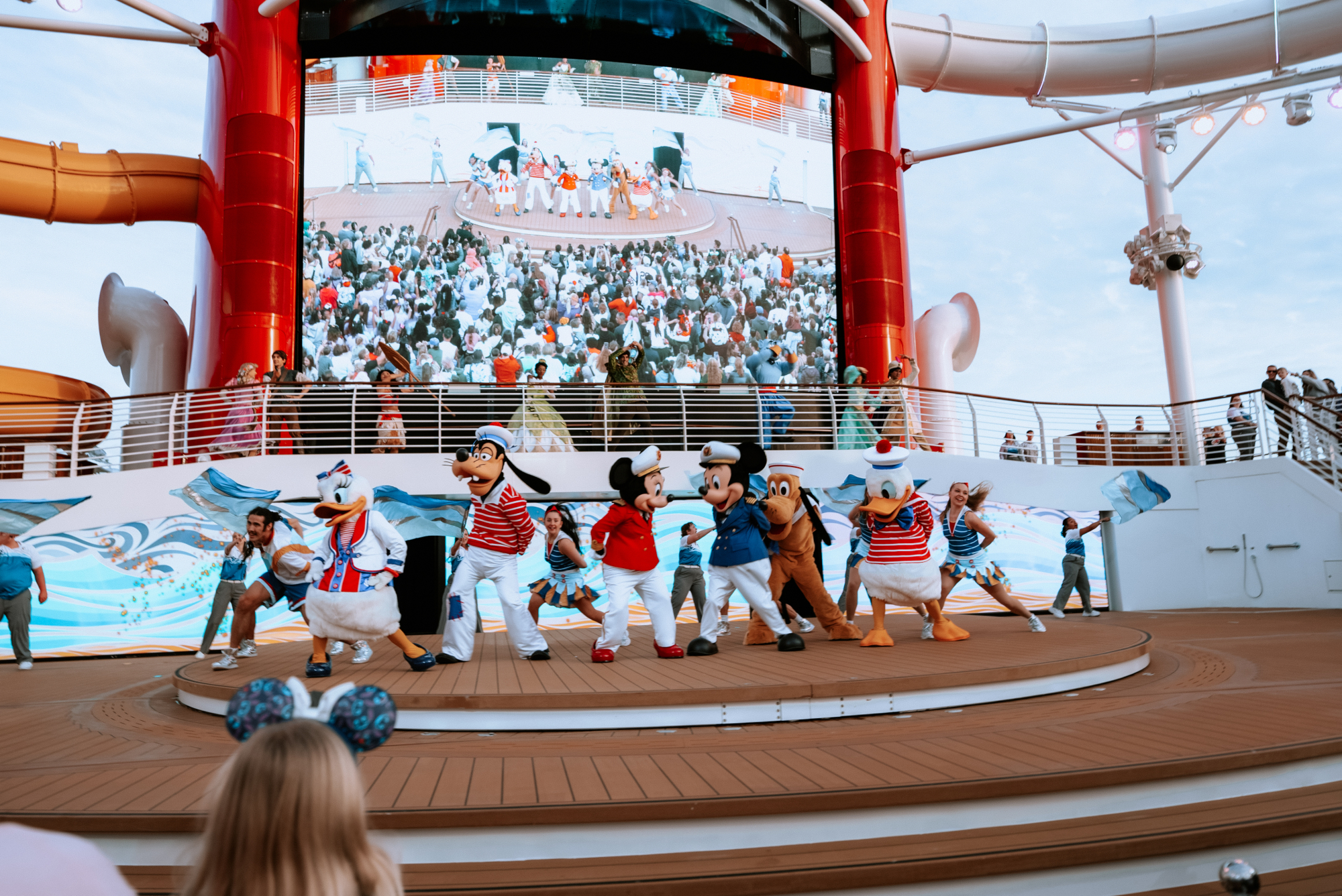 Disney characters perform on an outdoor cruise ship stage below a large screen