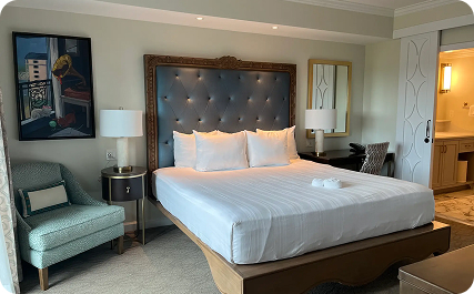 A Riviera Resort guest room has a tufted bed, white linens, a green chair, bedside lamps, and curtains