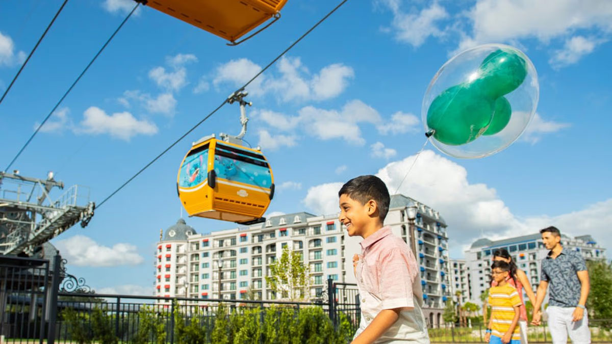 Guests walk below a yellow Disney Skyliner gondola beside a green Mickey-shaped balloon
