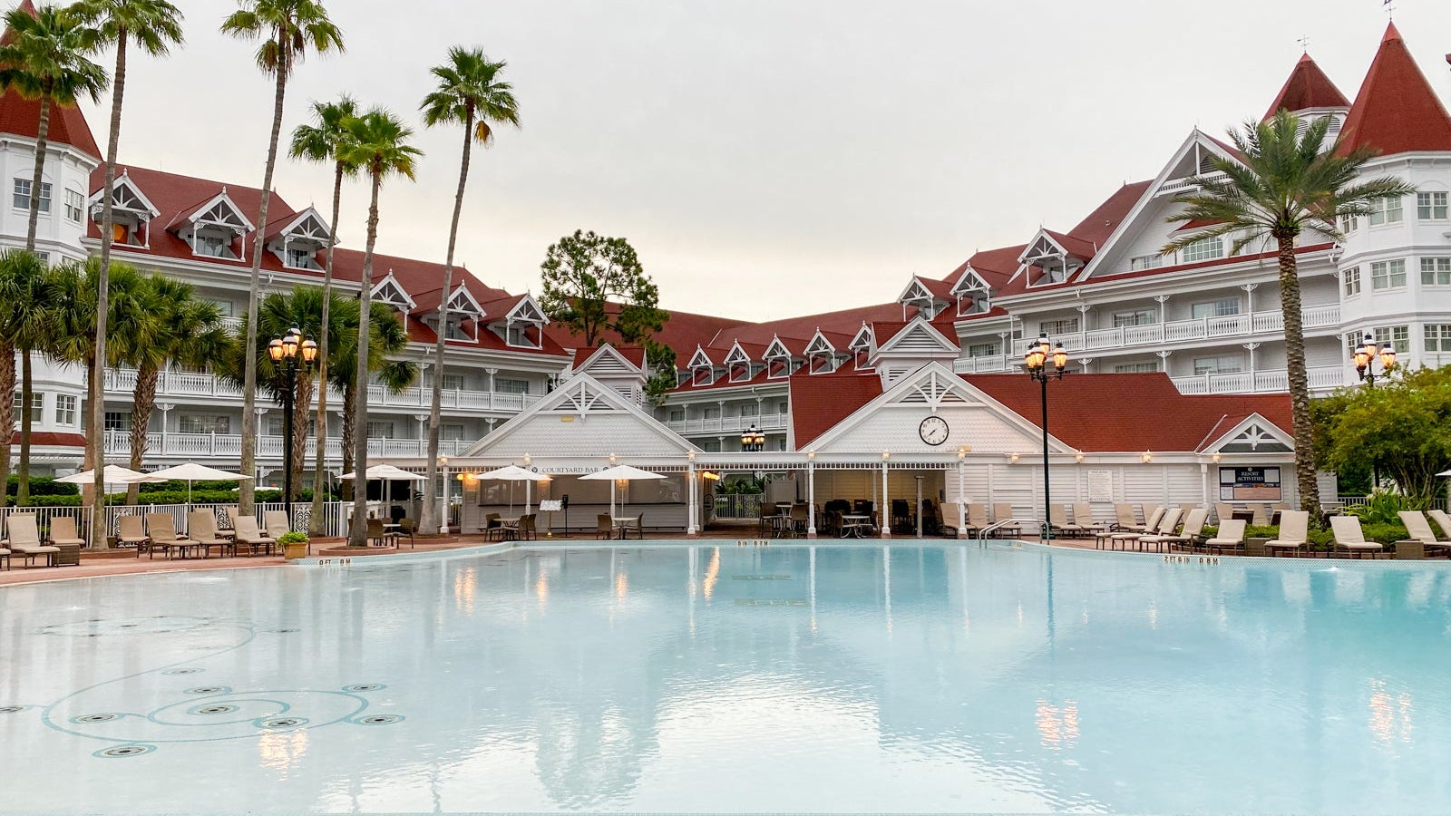 A pale blue swimming pool sits before white buildings with red roofs at the Grand Floridian