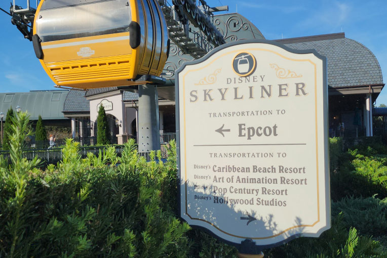 A yellow Skyliner gondola passes above an EPCOT direction sign
